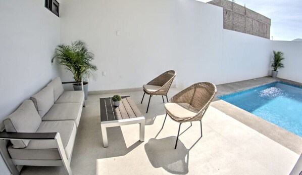 A rooftop patio with a pool, a sofa, two wicker chairs, and potted plants.