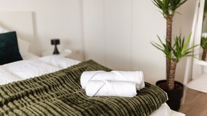 A neatly made bed with a green textured blanket and two rolled white towels, a potted plant, and a white chair in a room with white walls and a wooden floor. - Modern Stilvolle Fewo63qm 3 Zimmer (Hamm)