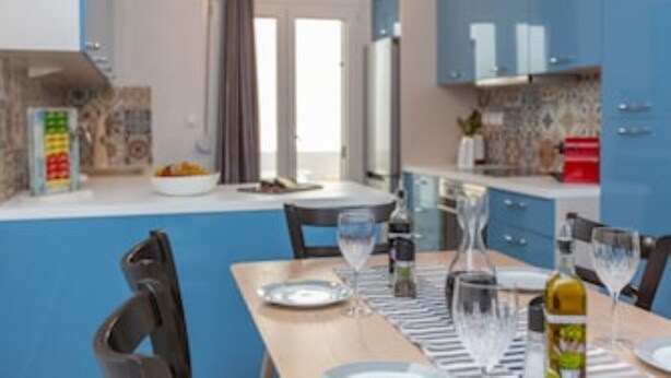 Una cocina moderna con armarios azules, una mesa de comedor preparada para dos personas y vista al área de estar a través de una puerta abierta.