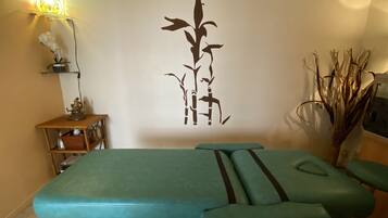Un cuarto de tratamientos con una camilla de masaje, una planta decorativa en la pared, un estante con una planta en maceta y una pequeña estatua.