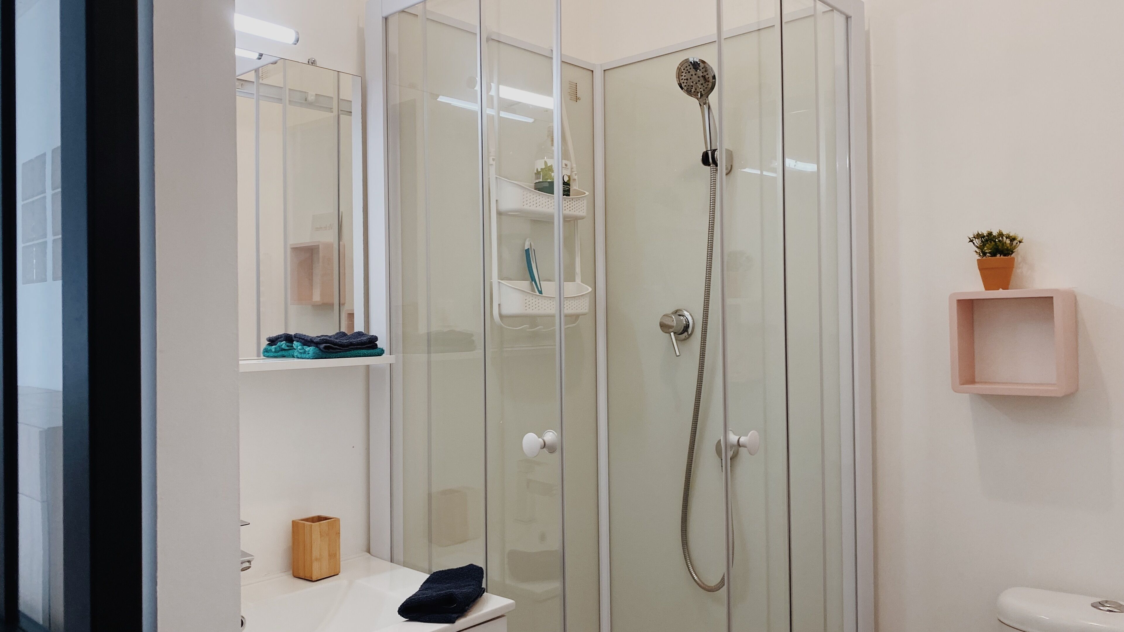 Une salle de bain moderne avec une douche à parois de verre, un lavabo blanc avec un récipient en bois et une petite étagère contenant des articles de toilette.