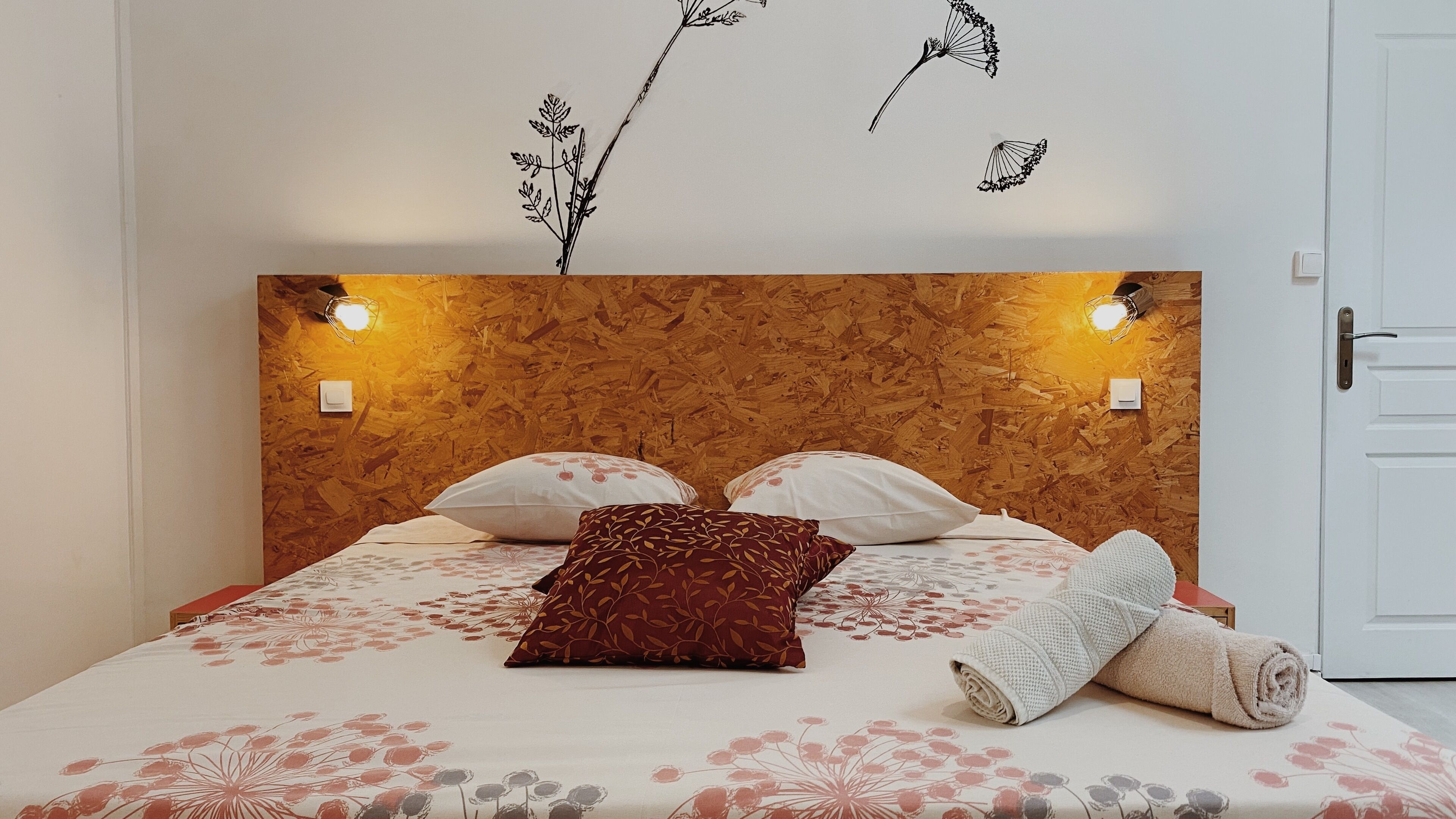 Een bed met bloemenbeddengoed, een houten hoofdbord, twee wandlampen en een deur rechts.