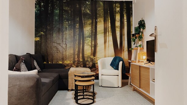 Un salón con un tapiz mural con motivos forestales, un sofá gris, un sillón blanco, una mesita de madera y un mueble bajo para televisión con una lámpara.
