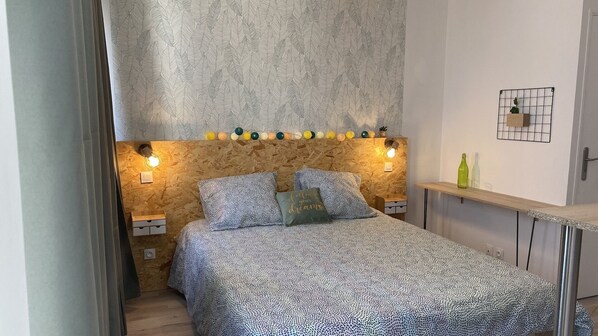 A bedroom with a patterned bedspread, two bedside lamps, a wall-mounted shelf with decorative items, and a door with a grid pattern. - La maison d'Angelique - Coliving Hotel - Gare de Niort (Niort)