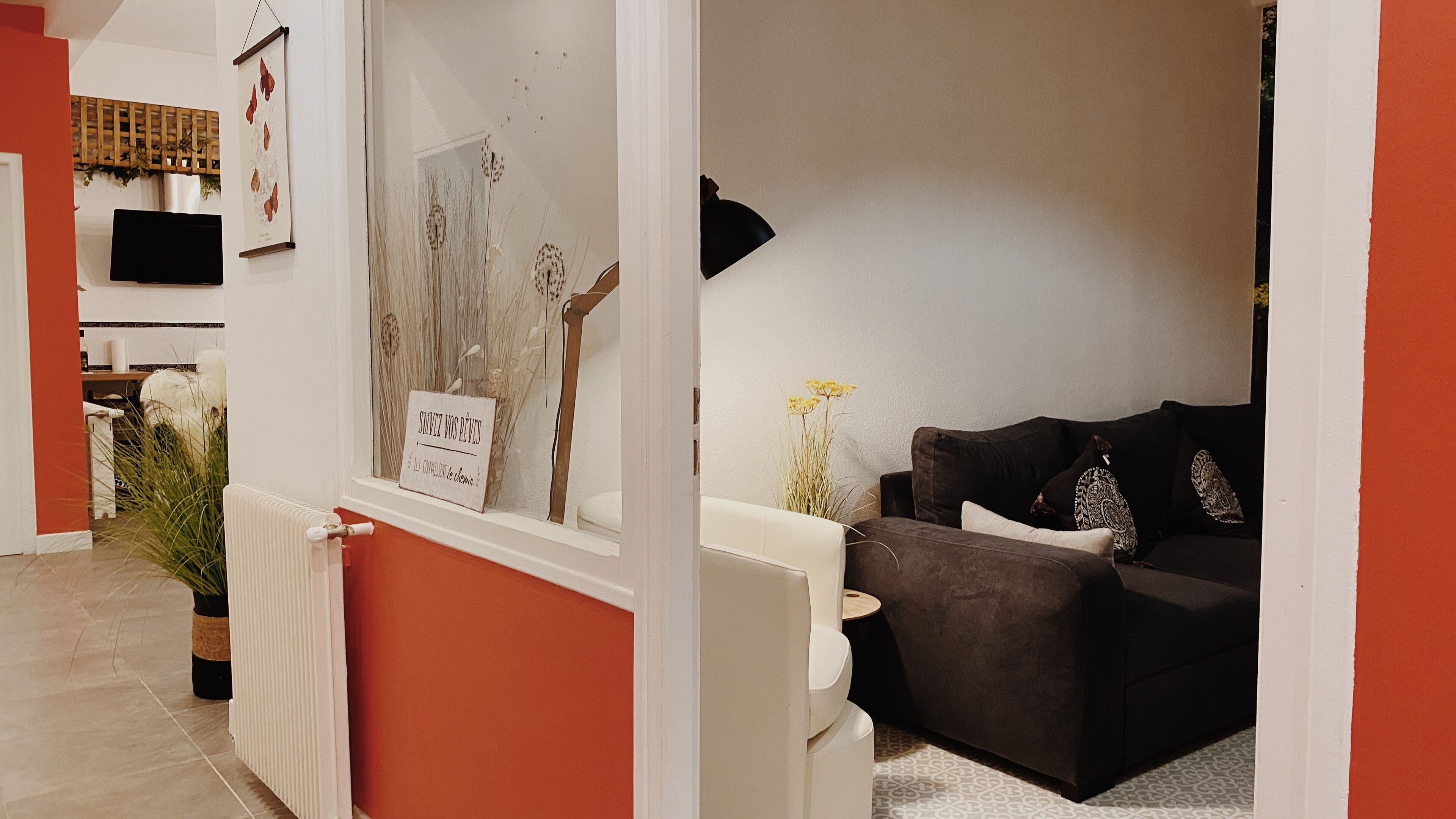 Een woonkamer met een donkerbruine bank, een witte fauteuil en een klein bijzettafeltje. Er is een raam met een bord waarop staat 