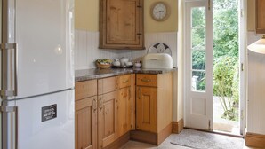 A kitchen with wooden cabinets, a white refrigerator, a clock, and a window with a view of greenery.