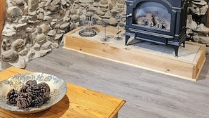Een stenen muur met een zwarte houtkachel, een houten tafel met een schaal vol dennenappels, en een plafondventilator.