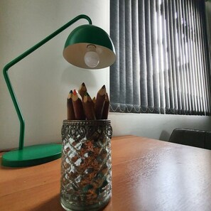 A desk with a green desk lamp, a glass mug with pencils, and a window with blinds.