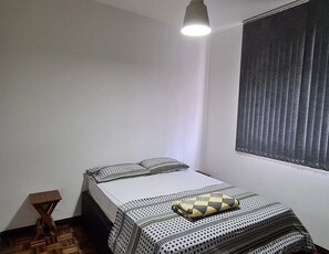 A small, neatly arranged bedroom with a single bed, a bedside table, and a window with blinds.