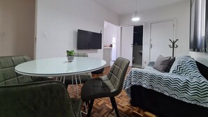 A modern living room with a round glass table, leather chairs, a sofa with a patterned throw, a flat-screen TV, and a coat rack. - Apto na Pampulha próximo ao Mineirão  (Belo Horizonte)