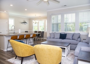 A modern kitchen with a central island, dining area with chairs, and a living room with a sofa and yellow chairs.