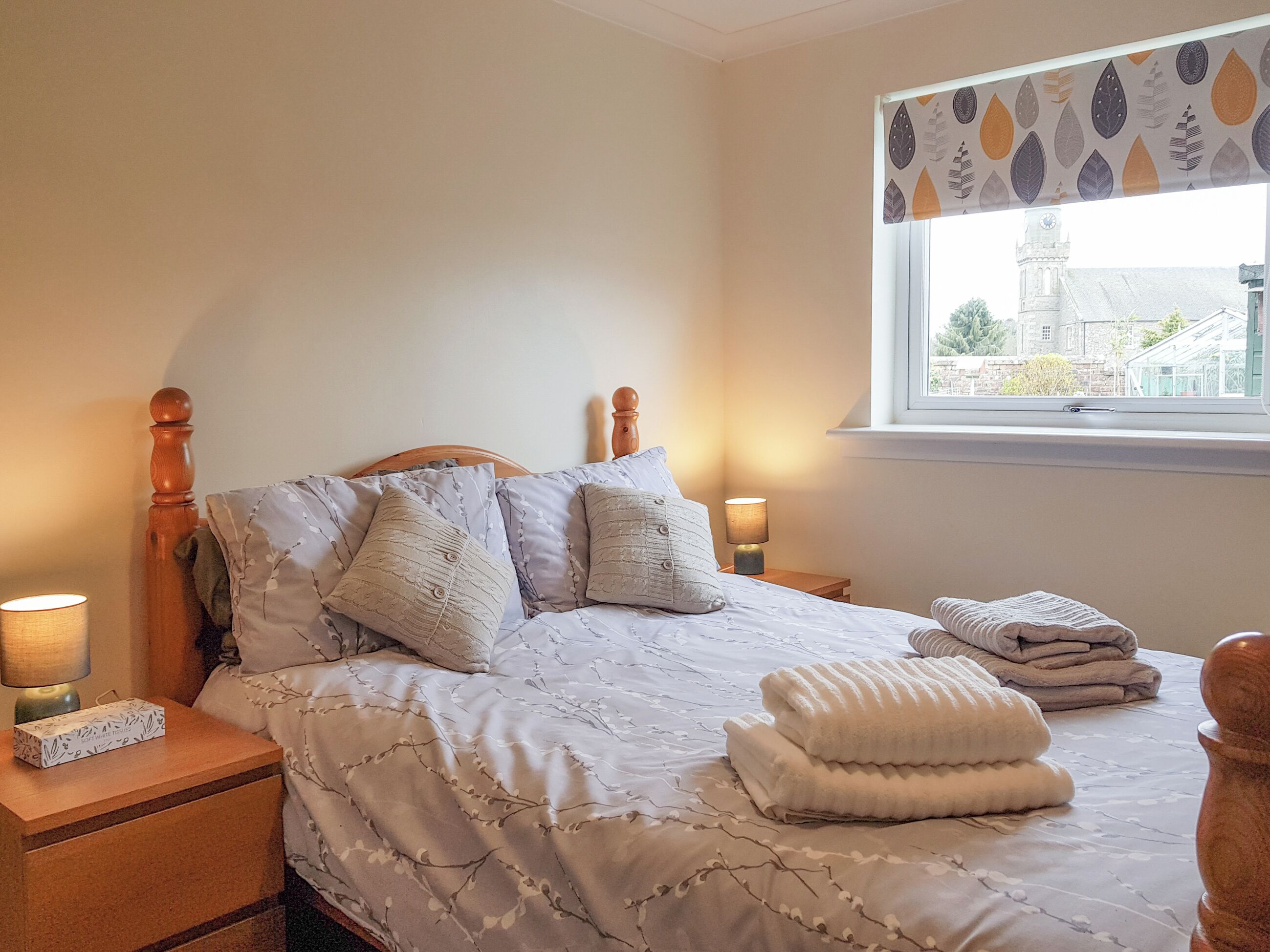 A neatly made bed with a floral-patterned duvet, two bedside lamps, and a window with a view of a building.