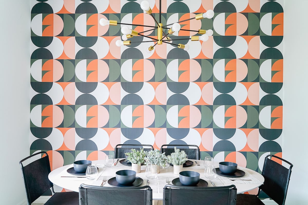 A dining area with a geometric patterned wallpaper, a round table set with black chairs, and a modern chandelier.