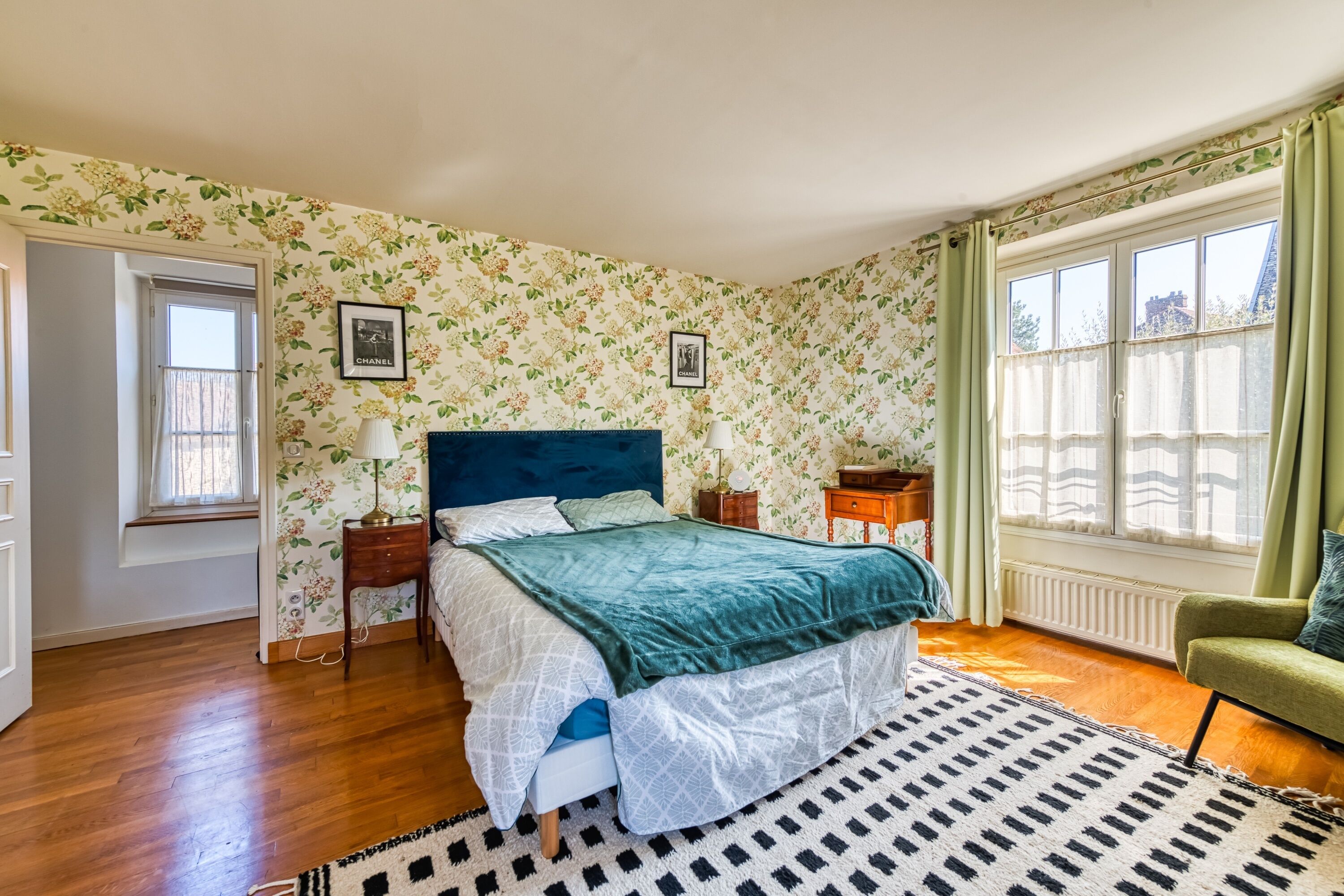 Un dormitorio con papel tapiz floral, una cama con cabecera azul, dos mesitas de noche con lámparas, un sillón verde y un ventanal con cortinas.