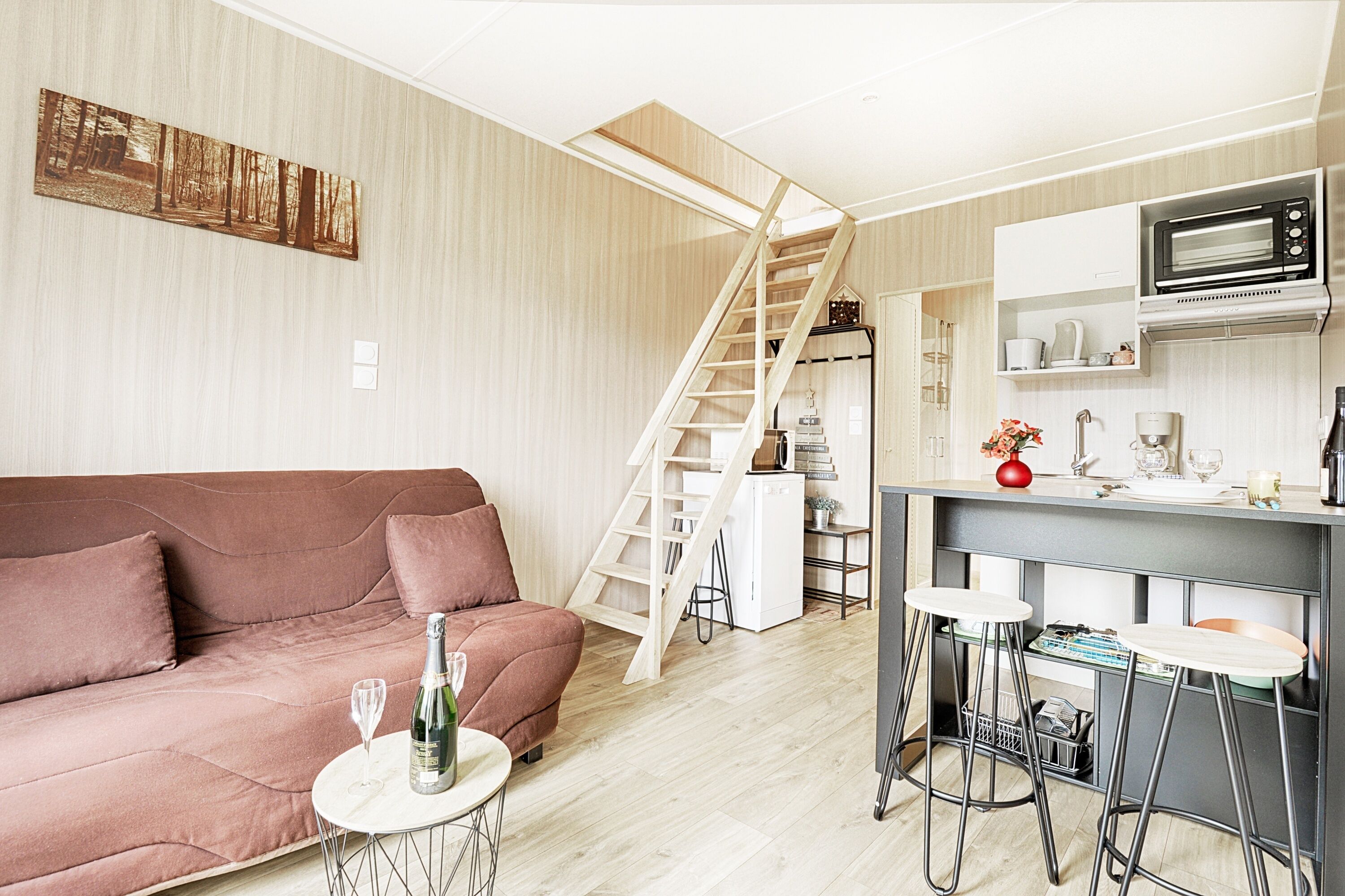 A modern living space with a sofa, a small table with a bottle and glass, a kitchen area with a microwave, and a staircase.
