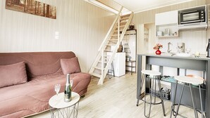 A modern living space with a sofa, a small table with a bottle and glass, a kitchen area with a microwave, and a staircase.