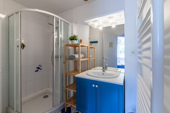 A bathroom with a glass-enclosed shower, a sink with a mirror, and a blue vanity with shelves. - La Grange - Charmante maison à Langrune-sur-mer (Langrune-sur-Mer)