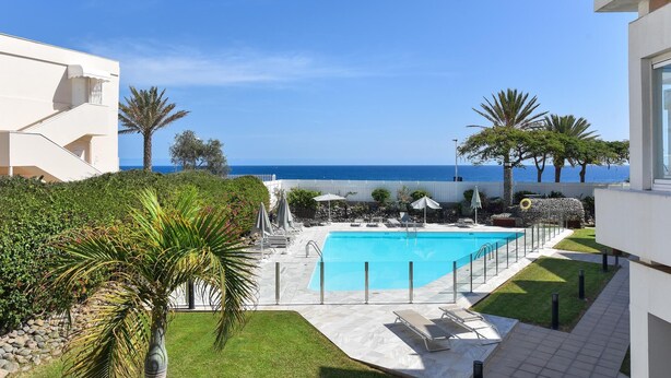 Zona de piscina con vista al mar, rodeada de palmeras y un edificio blanco.
