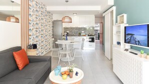 Una sala moderna con un sofá gris, un comedor con sillas blancas, una cocina con armarios blancos y un televisor sobre un mueble blanco.
