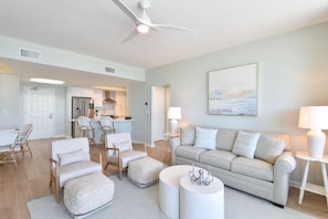 Una sala de estar moderna con un sofá color beige, sillones blancos y un comedor, con una cocina al fondo.
