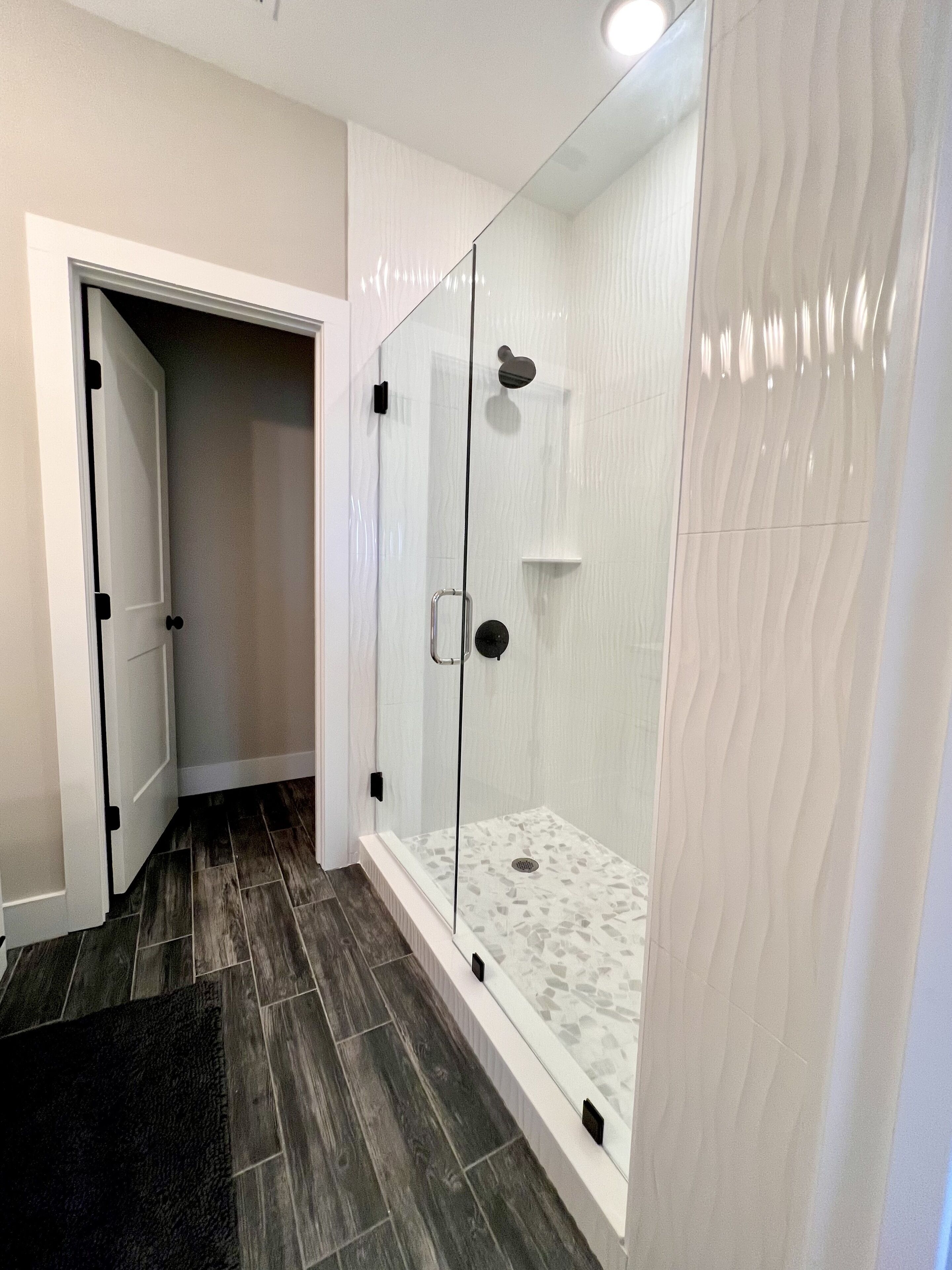 A modern bathroom with a glass-enclosed shower, dark wood flooring, and a white tiled wall.
