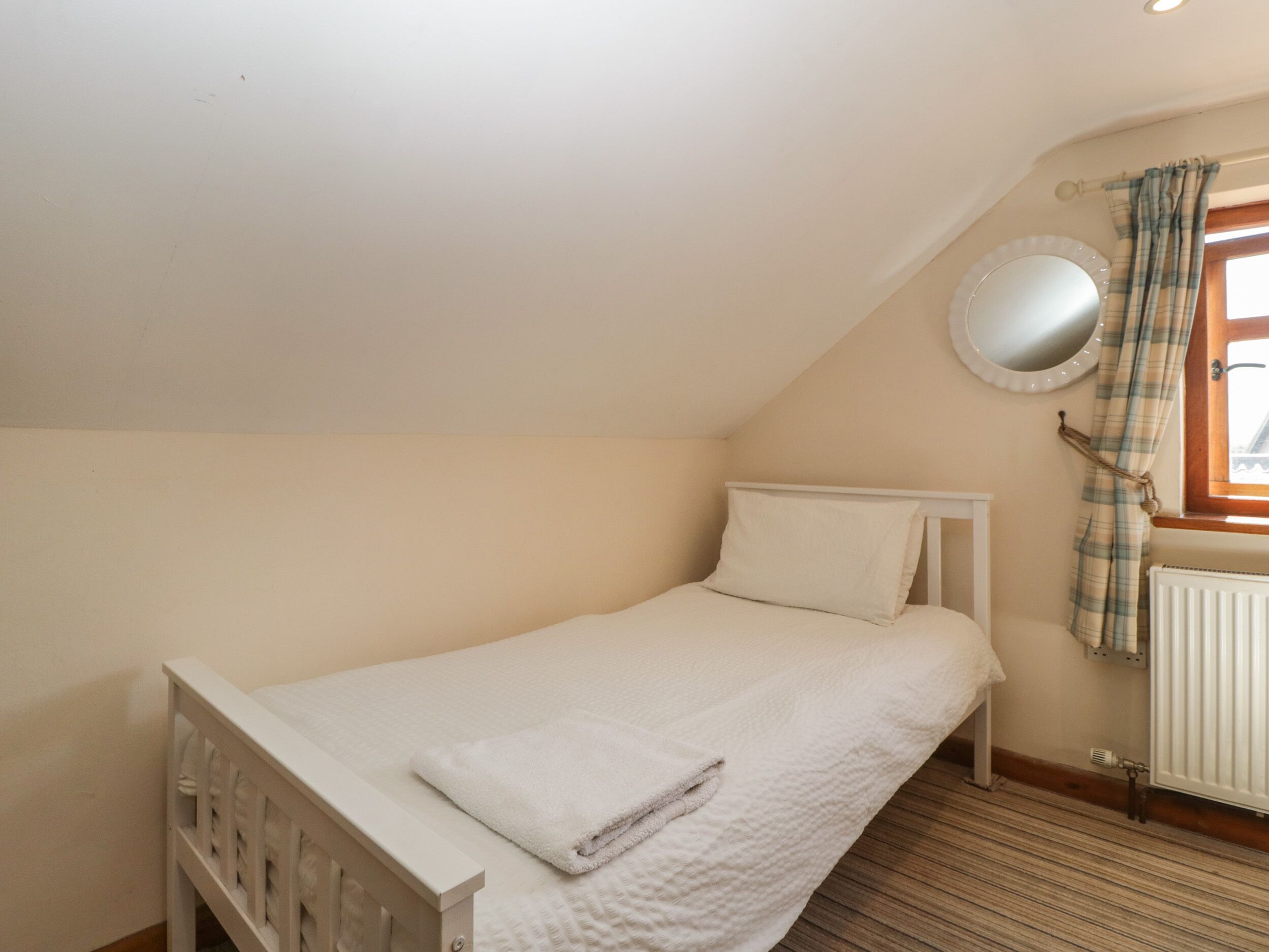 A small, single-bed room with a sloped ceiling, a window with plaid curtains, a round mirror, and a radiator.