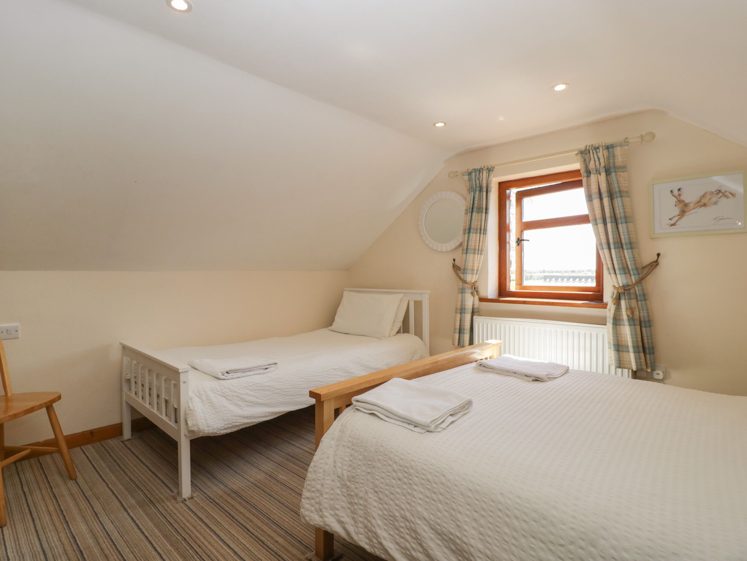 A bedroom with two single beds, a wooden bench, a radiator, a window with curtains, and a wall-mounted round mirror.