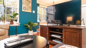 A living room with a large flat-screen TV, a sofa, a coffee table with a remote control, a potted plant, and a bookshelf.