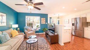 A living room with a sofa, coffee table, and TV, a kitchen with a refrigerator and cabinets, and a ceiling fan.