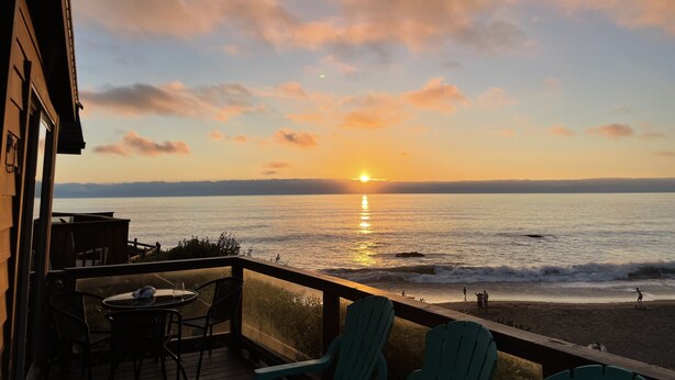 A balcony with a view of the sunset over the ocean, a table with chairs, and a person standing by the water. - Oceanfront Cabin 4 with Private Jacuzzi & Stunning Views (Smith River)