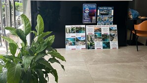 A reception desk with a glass partition, air conditioning unit, hanging pendant lights, and a refrigerator. There are promotional posters for day trips and a bottle of Erdinger Weissbier.