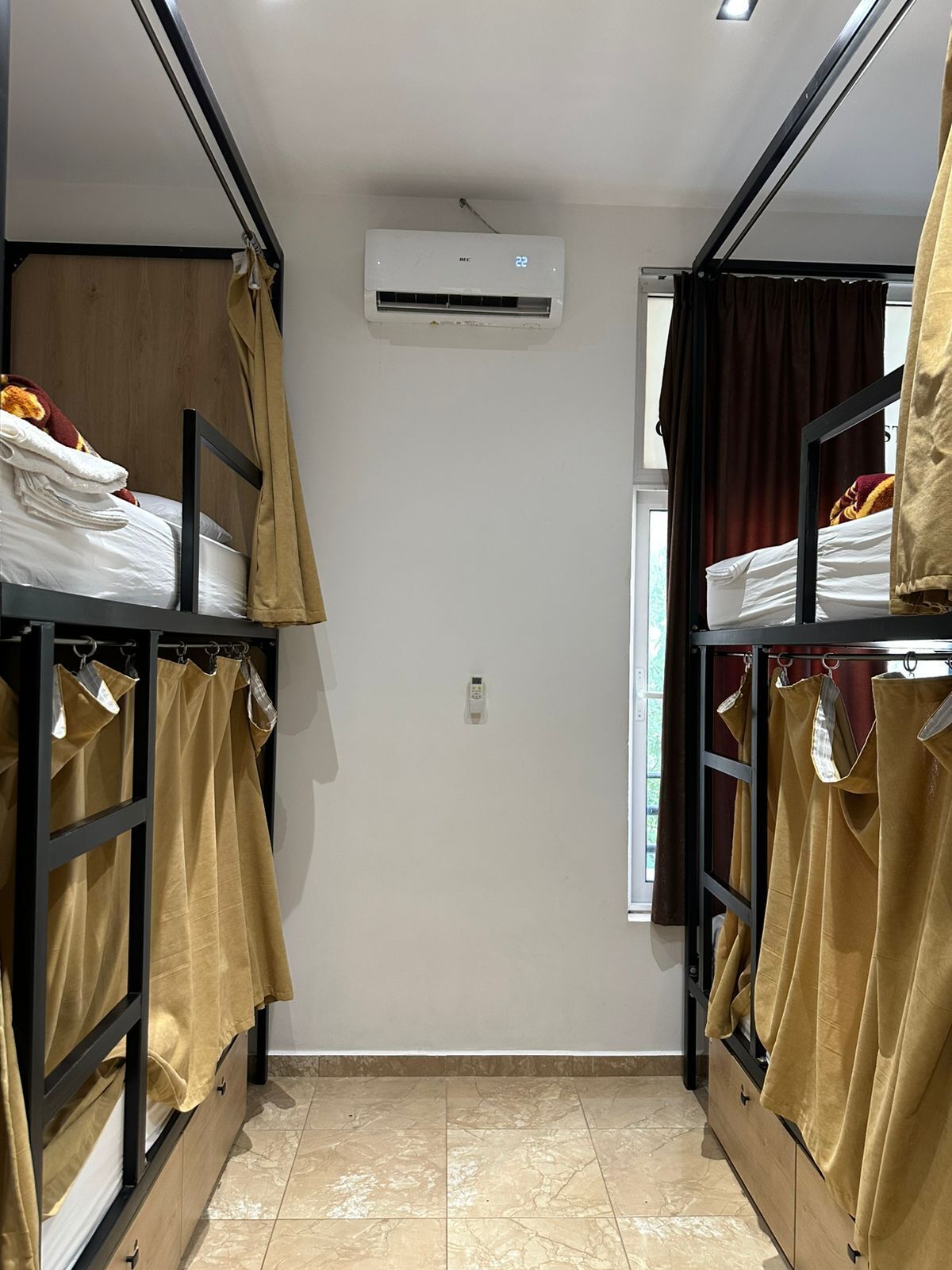 Habitación pequeña con literas, una ventana con cortinas, un equipo de aire acondicionado y un termostato en la pared.