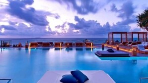 Zona de piscina con sillones de playa y sombrillas, vista despejada al océano y un cielo al atardecer.