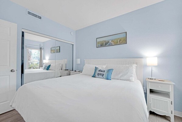 A bedroom with a large bed, a bedside table, a nightstand, a mirror, and a wall-mounted picture. - Quiet Destin Beachfront Resort! (Destin)