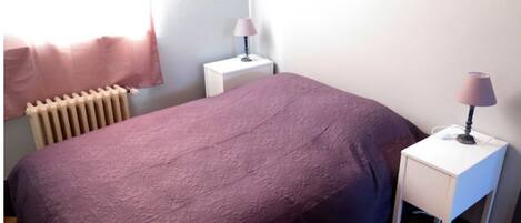 Une chambre à coucher avec un lit, une table de chevet avec une lampe, un radiateur et une fenêtre avec des rideaux.