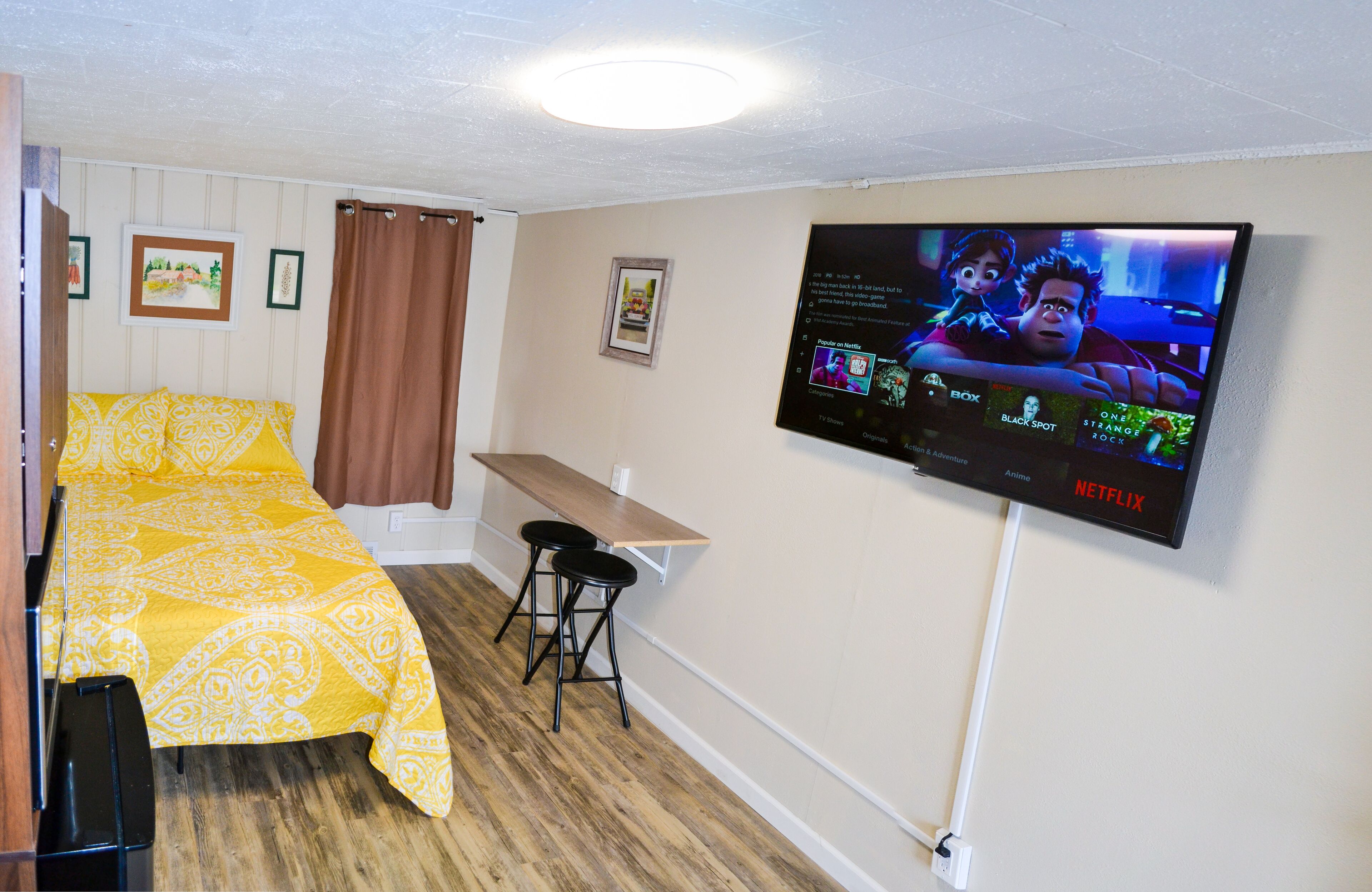 Une pièce comprenant un lit, un bureau, deux tabourets, un téléviseur fixé au mur et un luminaire au plafond.