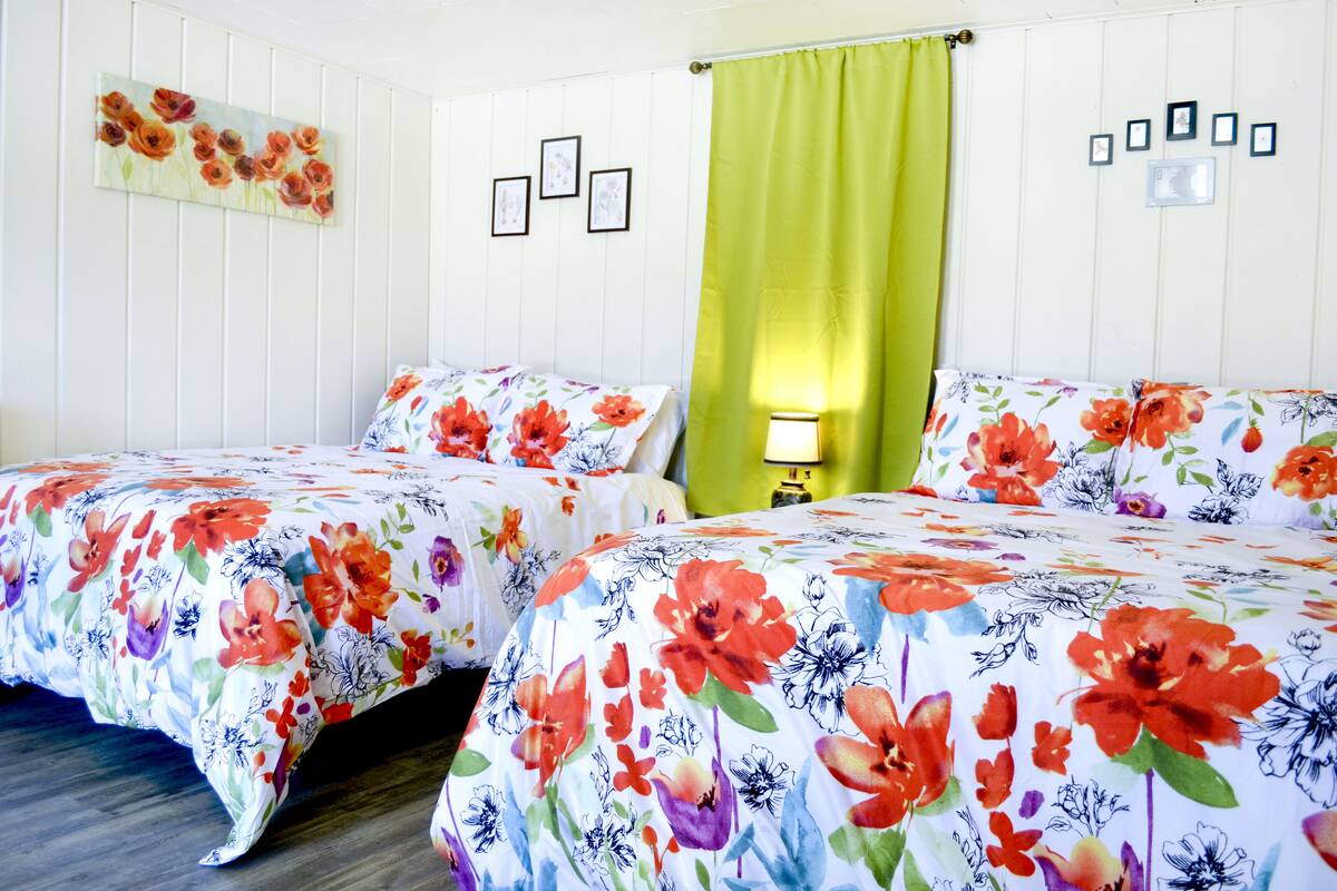 Une chambre d’hôtel avec deux lits, des couvre-lits à motifs floraux, un rideau vert et un mur orné de tableaux.