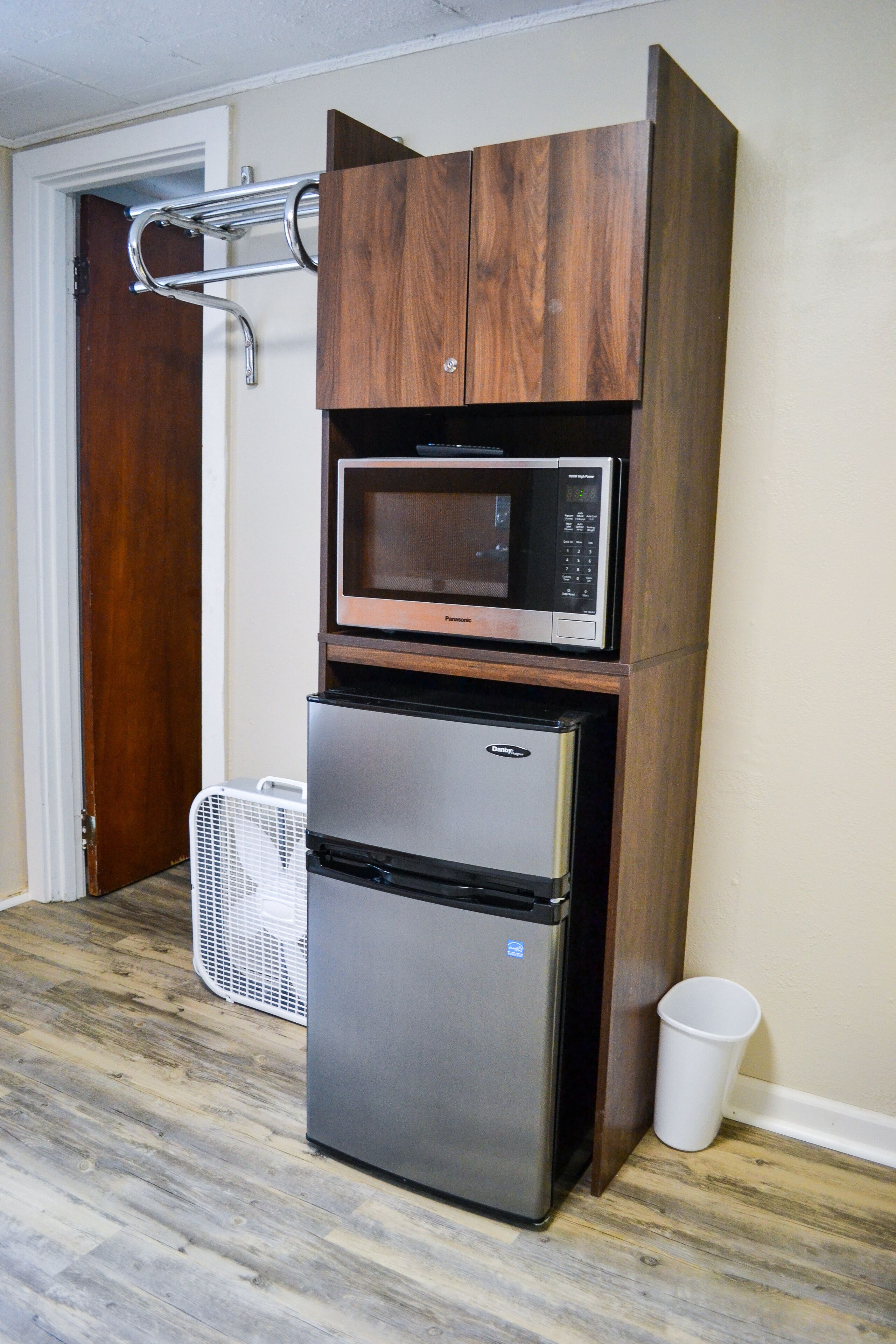 A compact kitchen with a built-in microwave and refrigerator, a small fan, and a trash bin.