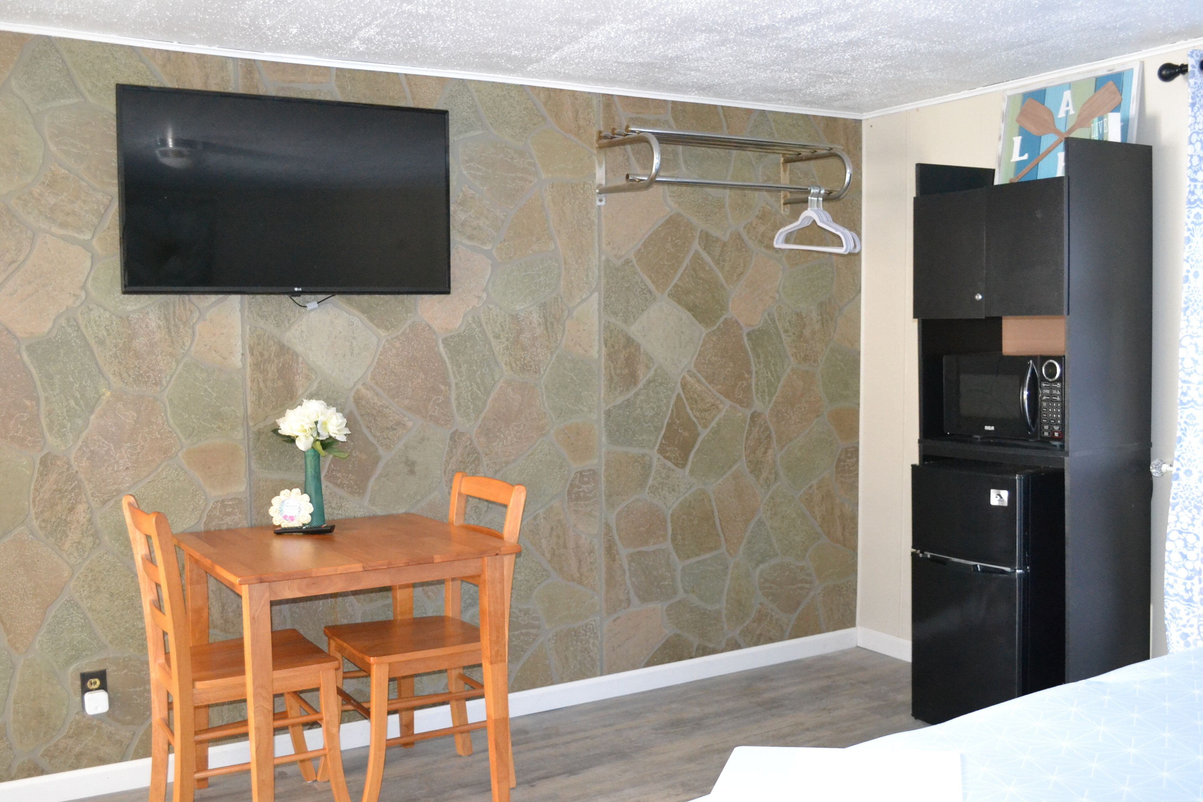 A room with a stone-patterned wall, a flat-screen TV mounted above a wooden table with chairs, a black refrigerator, and a microwave.