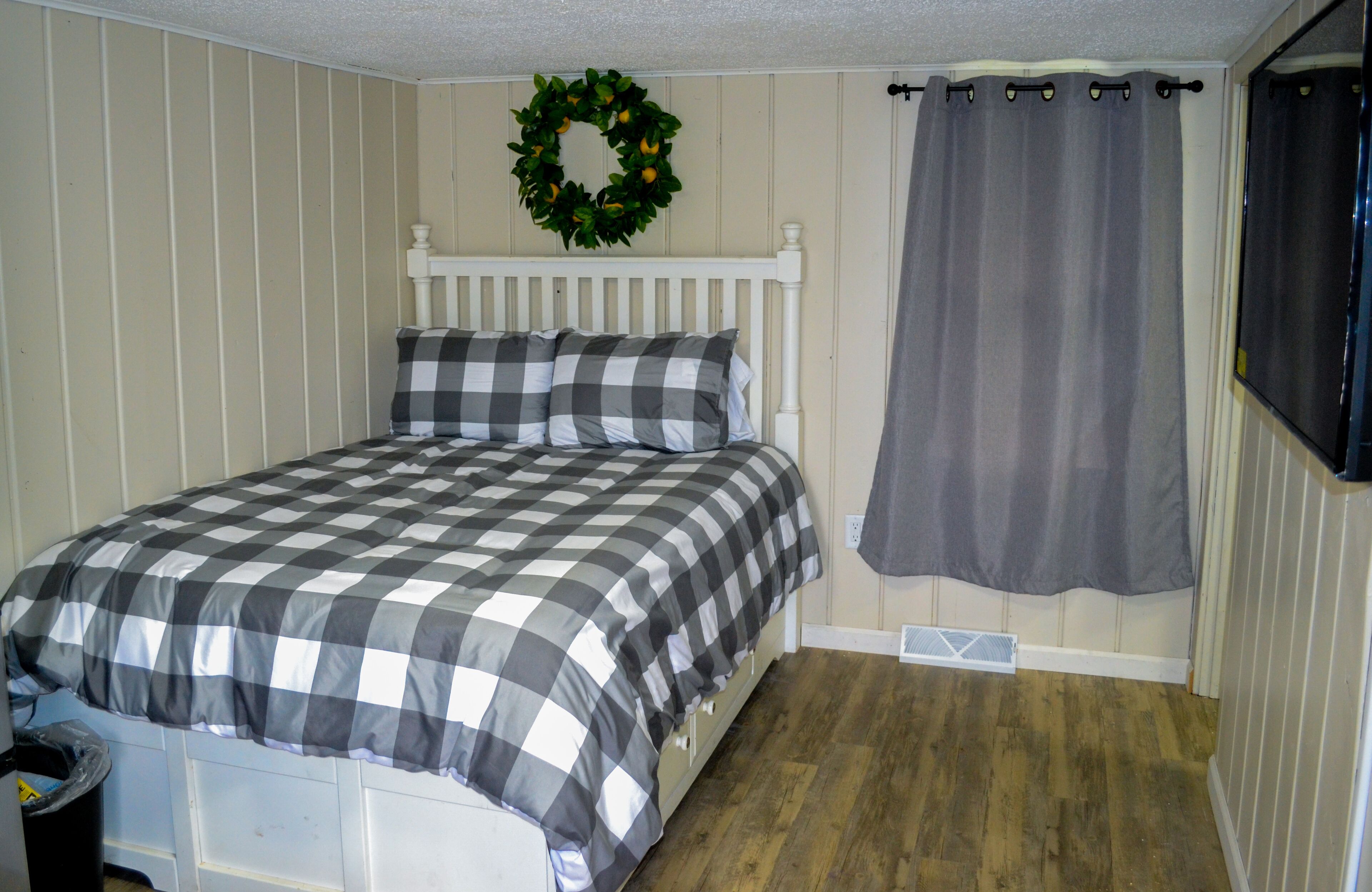 A bedroom with a checkered bedspread, a white headboard, a grey curtain, and a wooden floor.