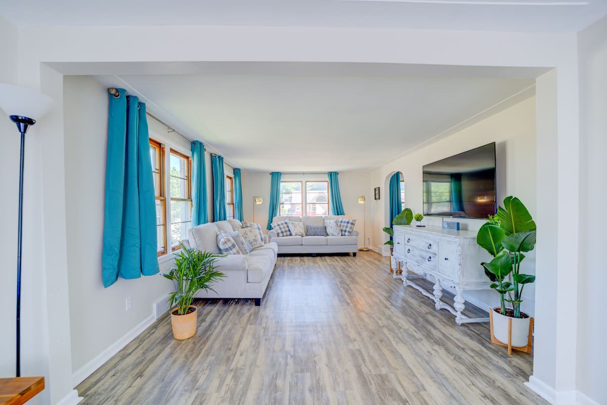 Un salon spacieux avec un canapé de couleur claire, une commode blanche et un téléviseur à écran plat. On y trouve également des plantes en pot et une lampe.