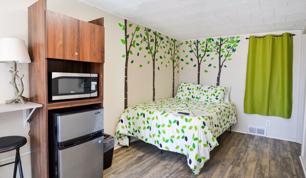 Habitación pequeña con cama, microondas, refrigerador y una pequeña mesa con lámpara.