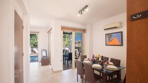 A dining area with a table set for four, a television on a stand, and a pool visible through the glass doors. - Villa Meena, 2BDR Kapparis Villa with Private Pool, Close to the Beach (Paralimni)
