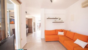 A modern living room with a large orange sofa, a flat-screen TV on a stand, and a dining area in the background. - Villa Meena, 2BDR Kapparis Villa with Private Pool, Close to the Beach (Paralimni)