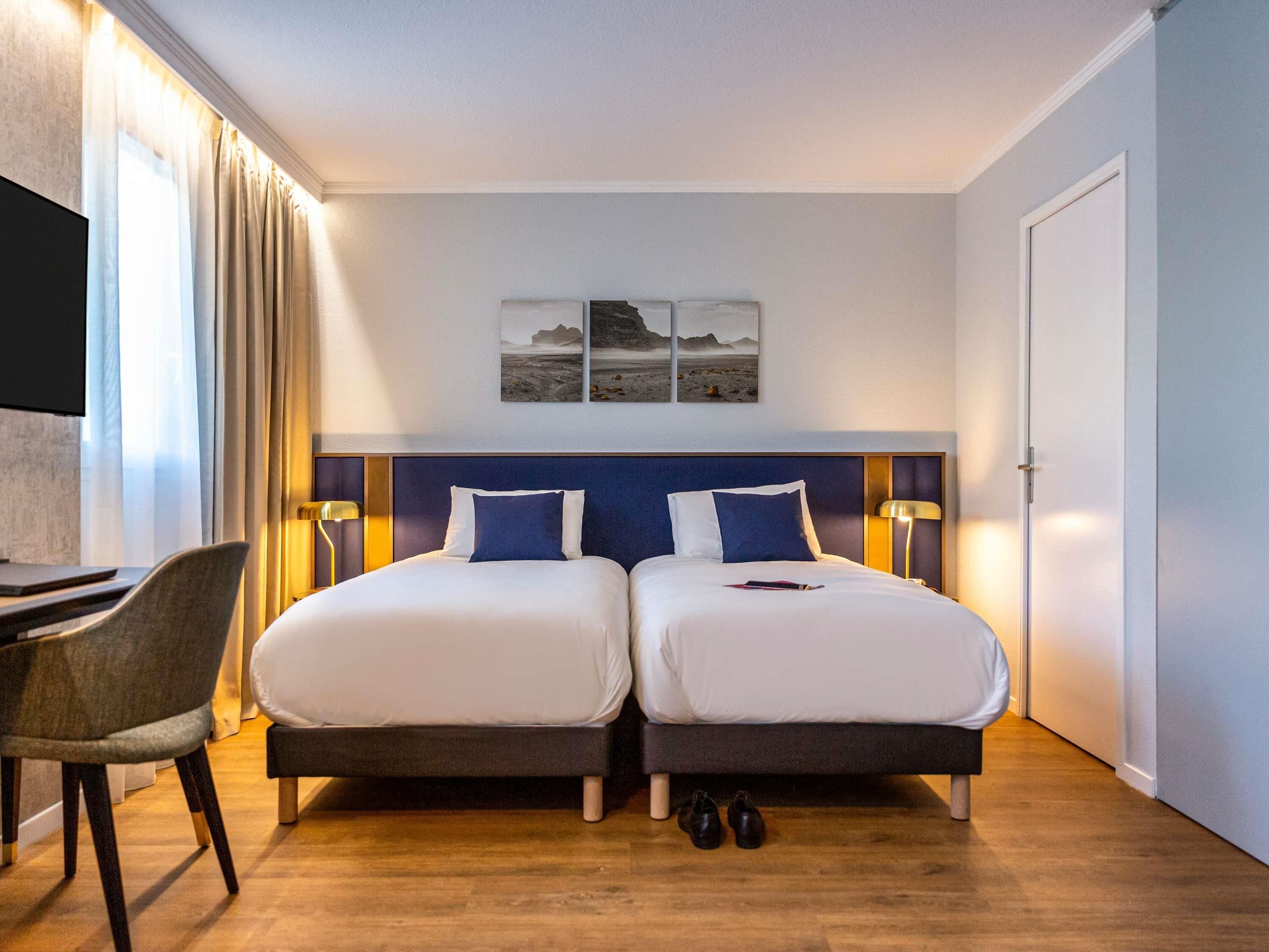 A hotel room with two beds, a desk, a chair, and a TV.