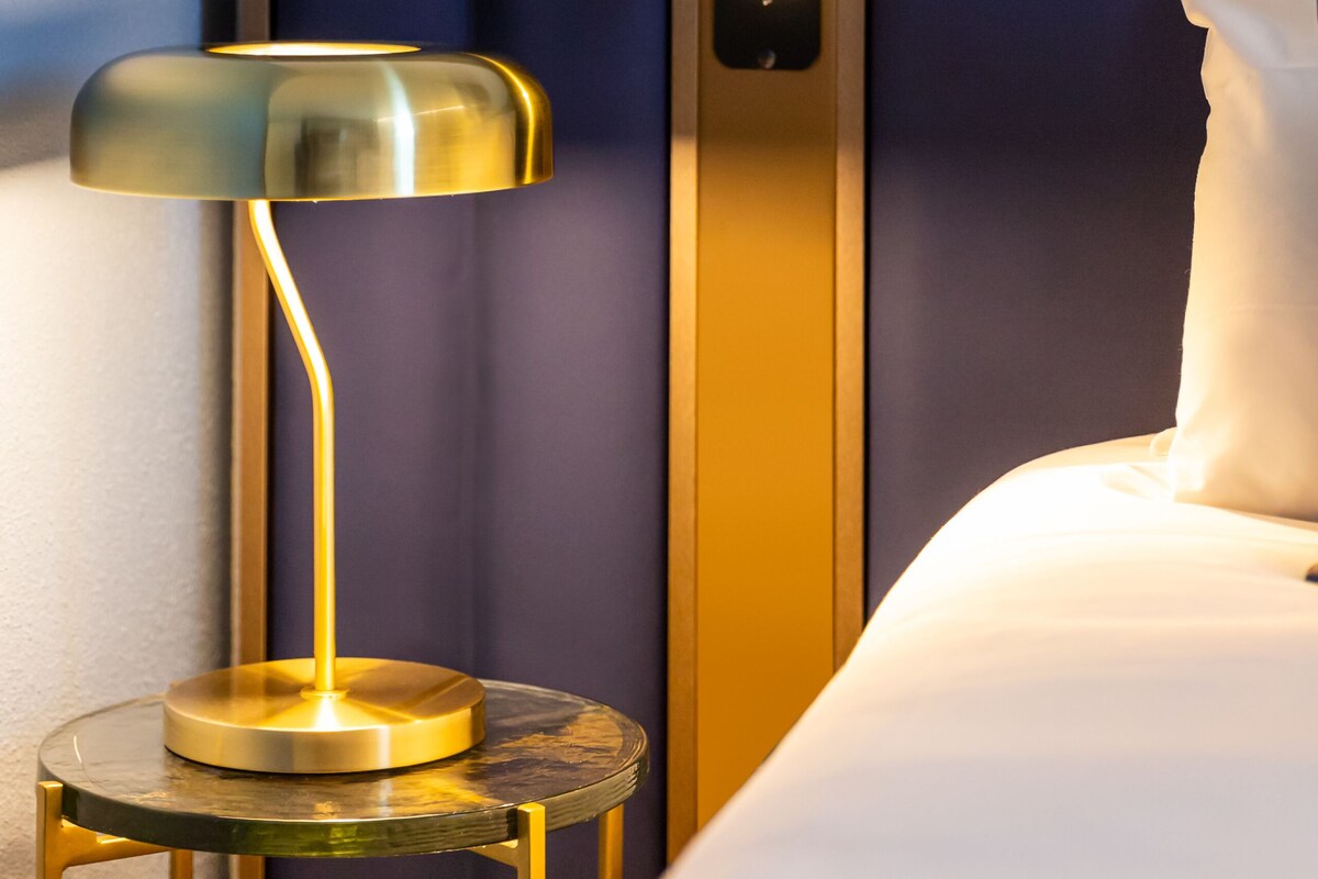 Una habitación de hotel con una cama, una mesita de noche con lámpara y un panel de control en la pared.