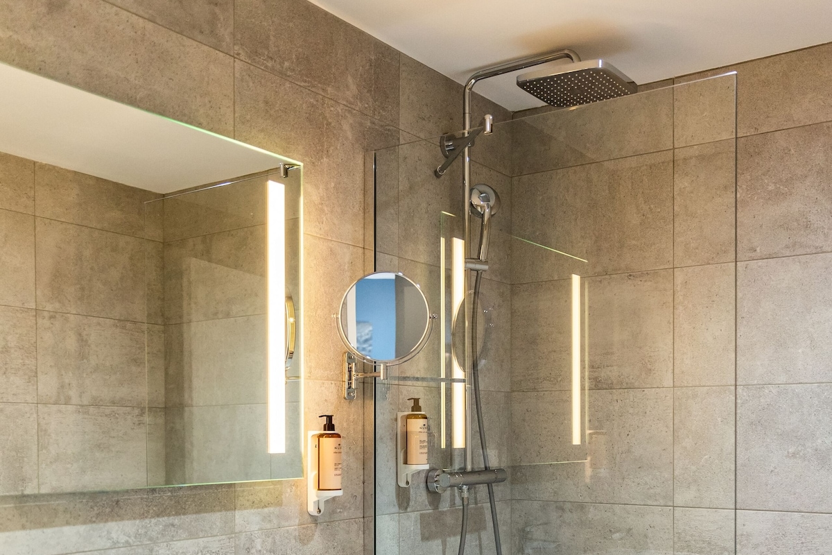 Un baño moderno con un espejo grande, un lavamanos blanco, una cabina de ducha de vidrio y piso de madera.