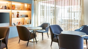 A modern room with a wooden floor, blue walls, a bookshelf with books and decorative items, a round table with chairs, and a large window with sheer curtains. - Mercure Toulouse Aéroport Blagnac (Blagnac)