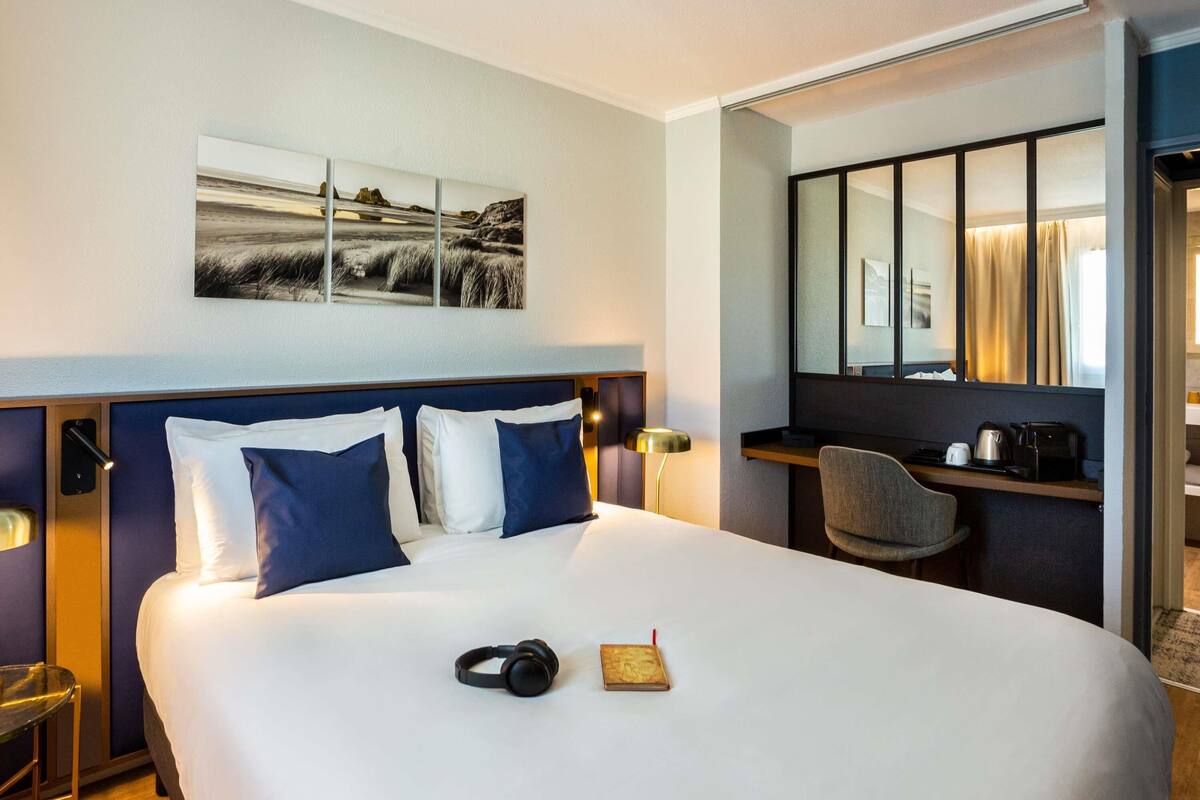 A hotel room with a large bed, a desk with a chair, a mirror, and a painting on the wall.