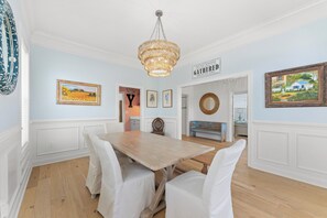 Comedor con una mesa de madera y sillas blancas, una araña y obras de arte en la pared.